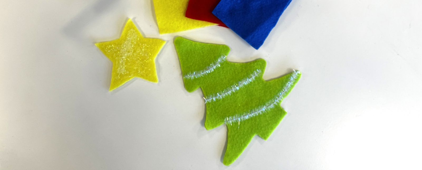 Julpyssel av filt, utskurna former av en gul stjärna och en grön julgran med glitterdetaljer, tillsammans med färgade filtark i rött, blått och gult på vitt underlag.